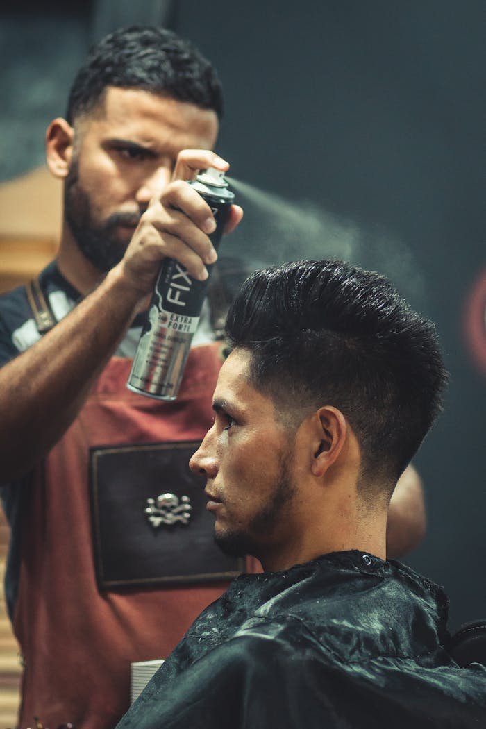Experienced stylist applies finishing spray to clients hair during grooming session at modern barbershop.